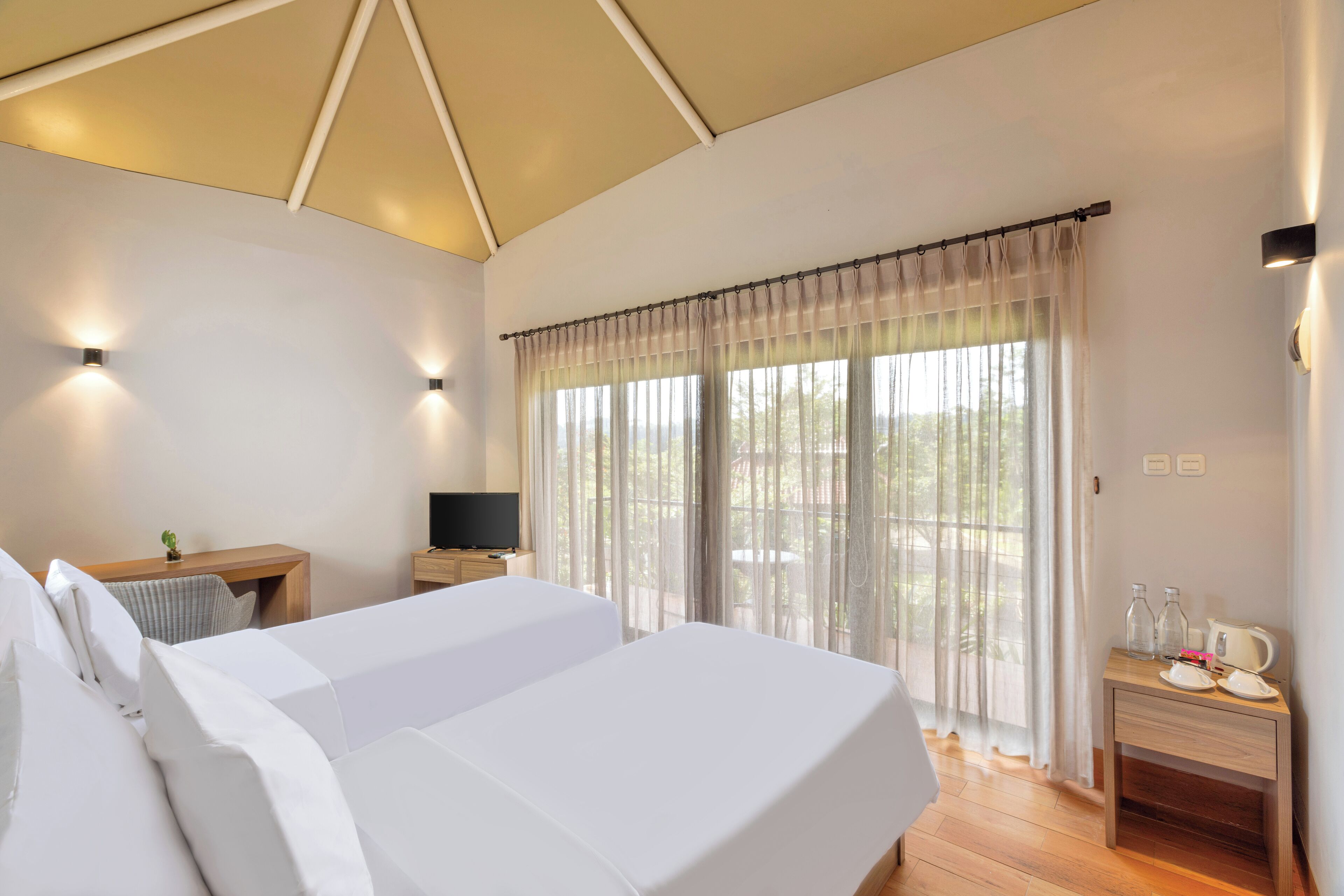 A hotel room with a large bed, a desk with a television, and a window with sheer curtains.