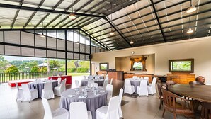 A spacious indoor area with round tables and chairs, a long wooden table, and a view of greenery outside. - The Glamping @ARTOTEL Pelangi Park (Bogor)