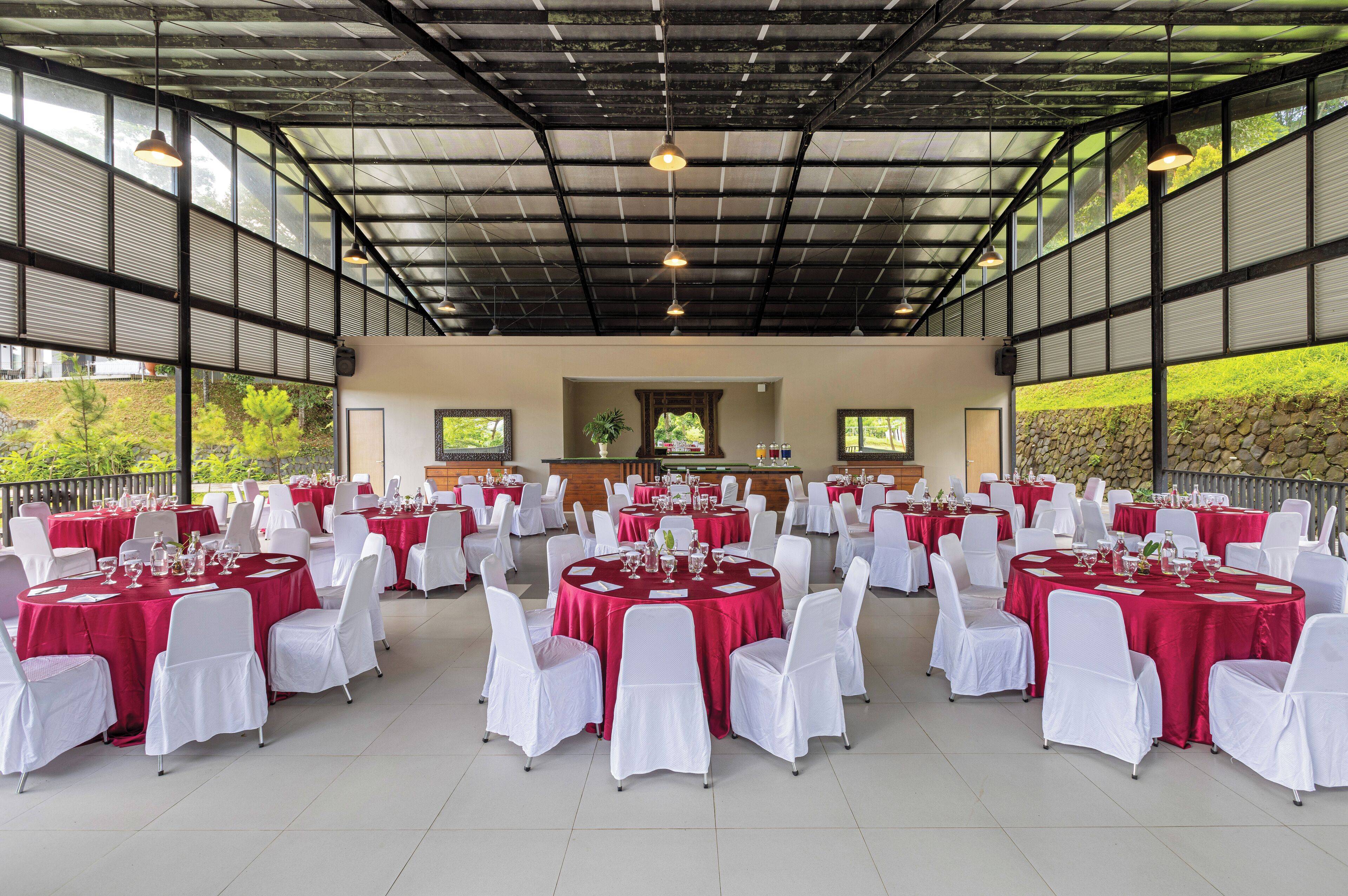 A spacious indoor venue with round tables covered in red tablecloths and white chairs, featuring a high ceiling with metal beams and large windows.