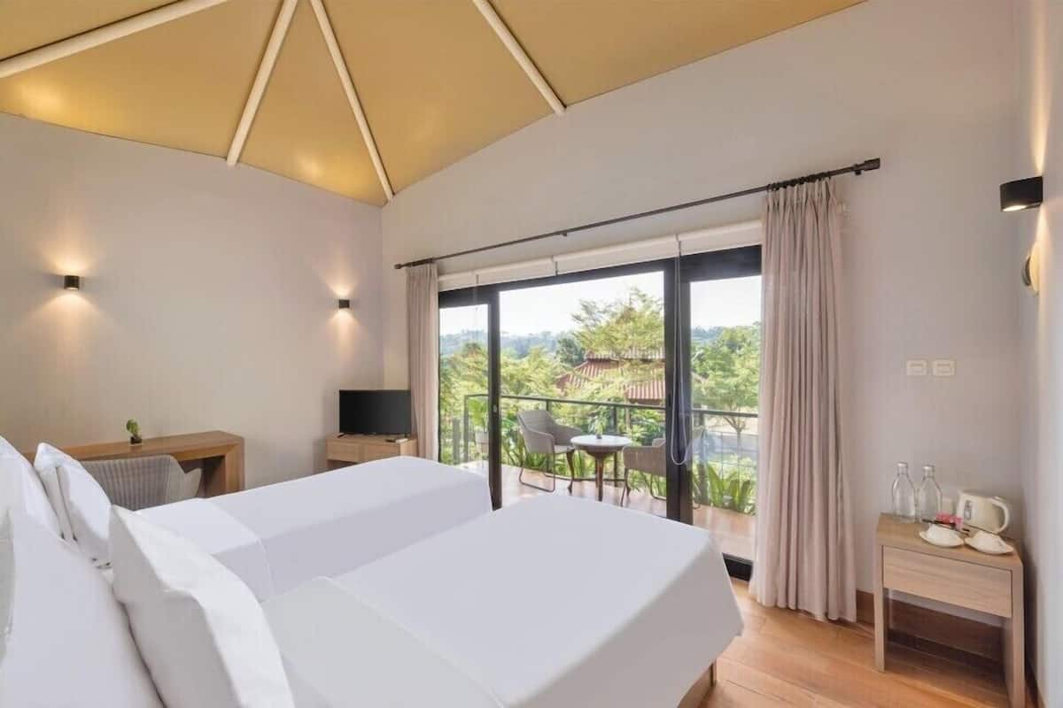 A modern hotel room with a large bed, a TV, a desk, and a balcony with a view of greenery.