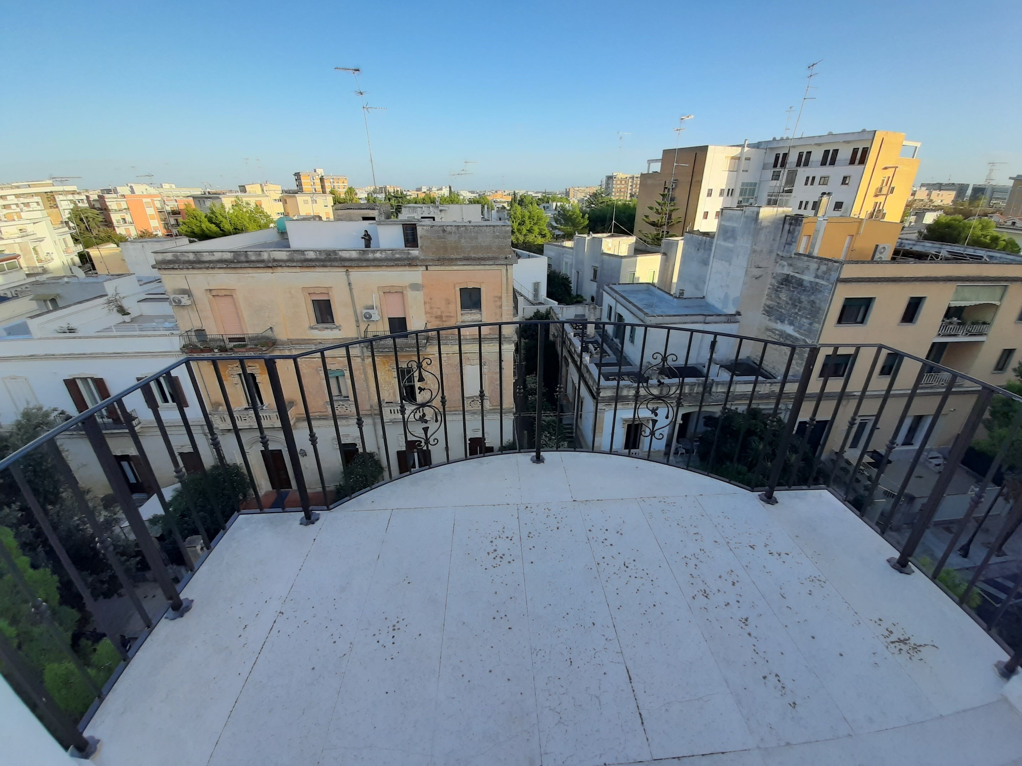 A rooftop terrace with a curved railing, offering a view of surrounding buildings and a clear sky.