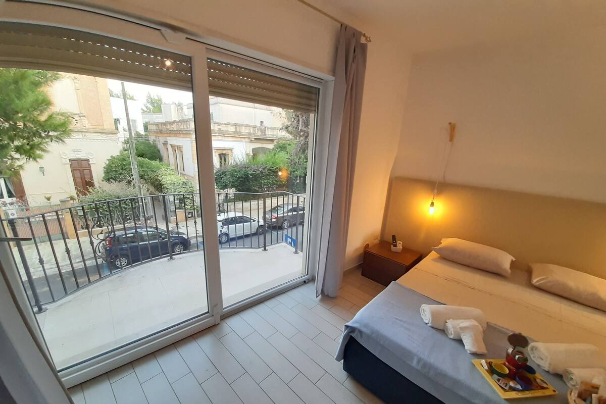 A hotel room with a balcony, a bed with pillows and towels, a bedside table with a lamp, and a view of the street and buildings outside.