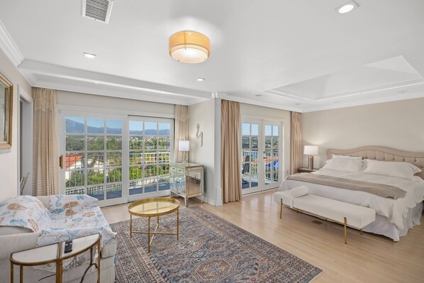 A spacious bedroom with a large bed, a sitting area with a small table, and a view of the city through large windows.