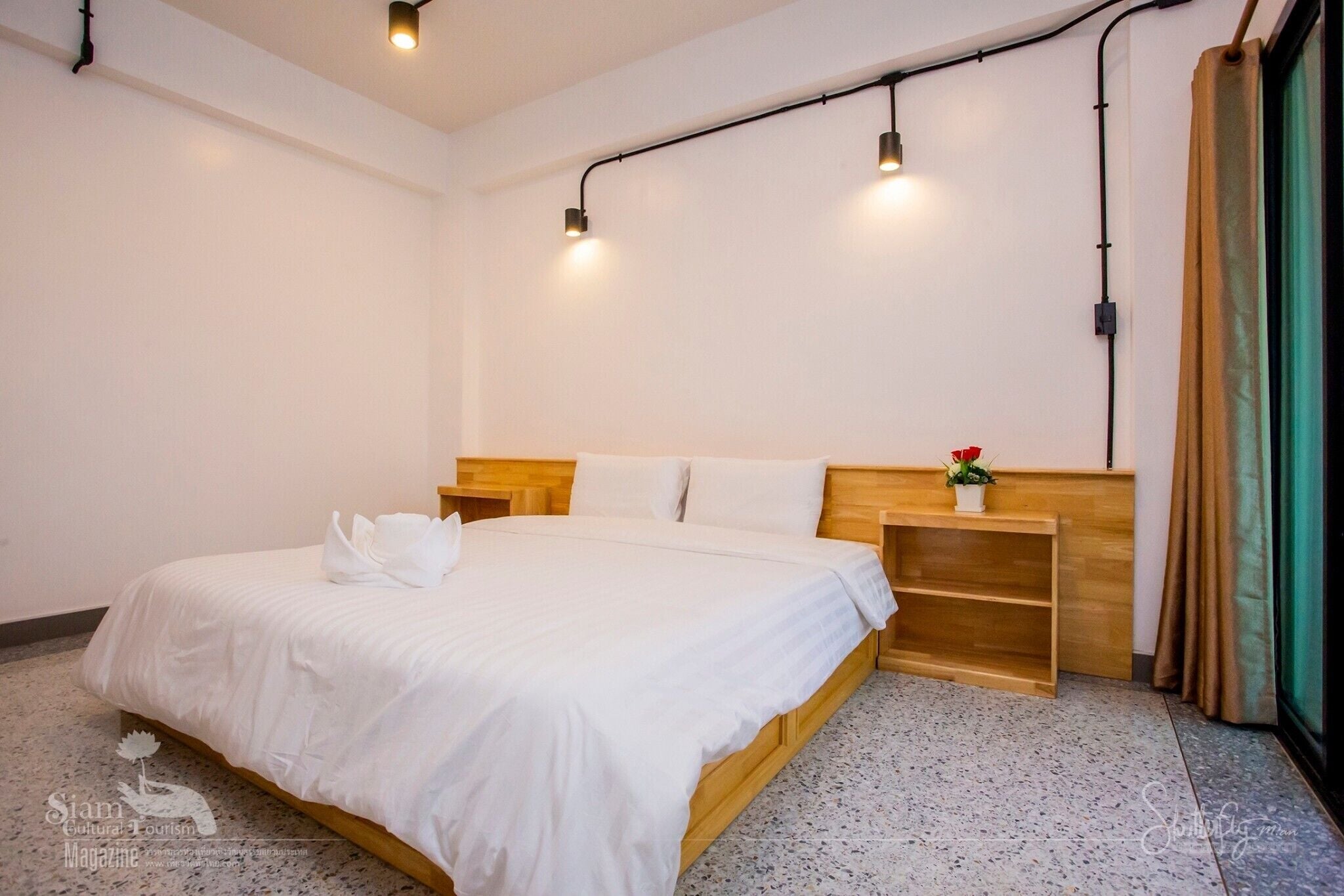 Ein ordentlich bezogenes Bett in einem Hotelzimmer, mit einem Holzkopfteil, einem kleinen Nachttisch mit einer Topfpflanze und einem Vorhangfenster.