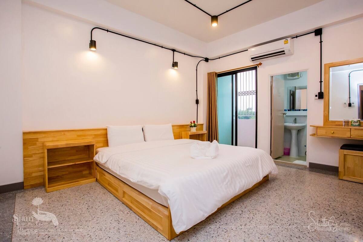 A hotel room with a wooden bed, white bedding, a small bedside table, a mirror, and an air conditioning unit.