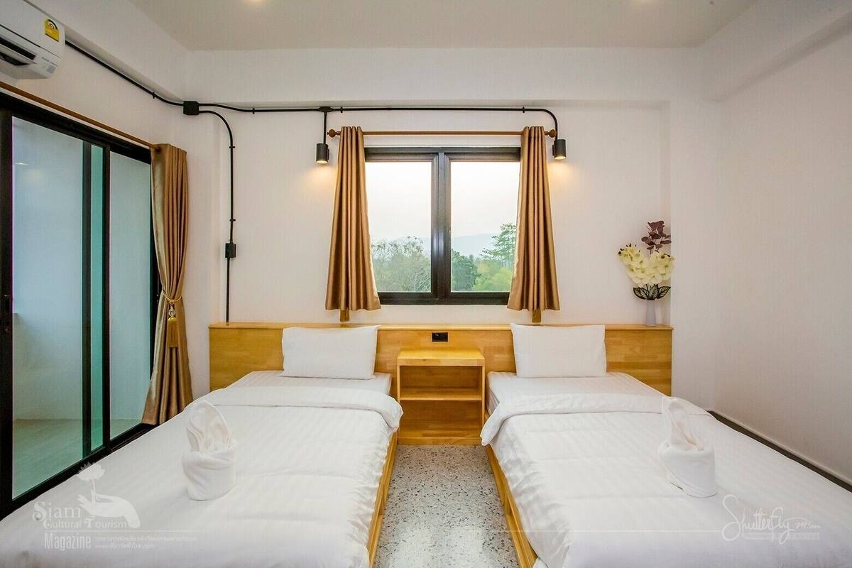 A hotel room with two single beds, a window with curtains, a wooden headboard, a small table, and a view of greenery outside.