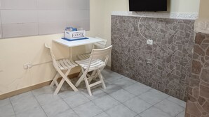 Een kleine kamer met een wit bureau, een stoel, een aan de muur bevestigde televisie en een muur met een steenmotief.