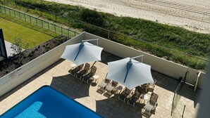 Un espace piscine avec des chaises longues et des parasols, donnant sur une plage où des gens se promènent.