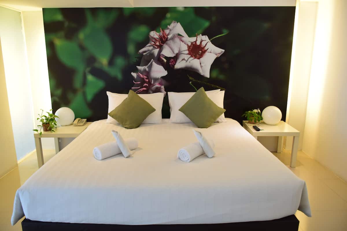 Un lit bien fait, avec des serviettes pliées, une table de chevet sur laquelle se trouvent un téléphone et une plante, et un mur orné d’un tableau représentant des fleurs.