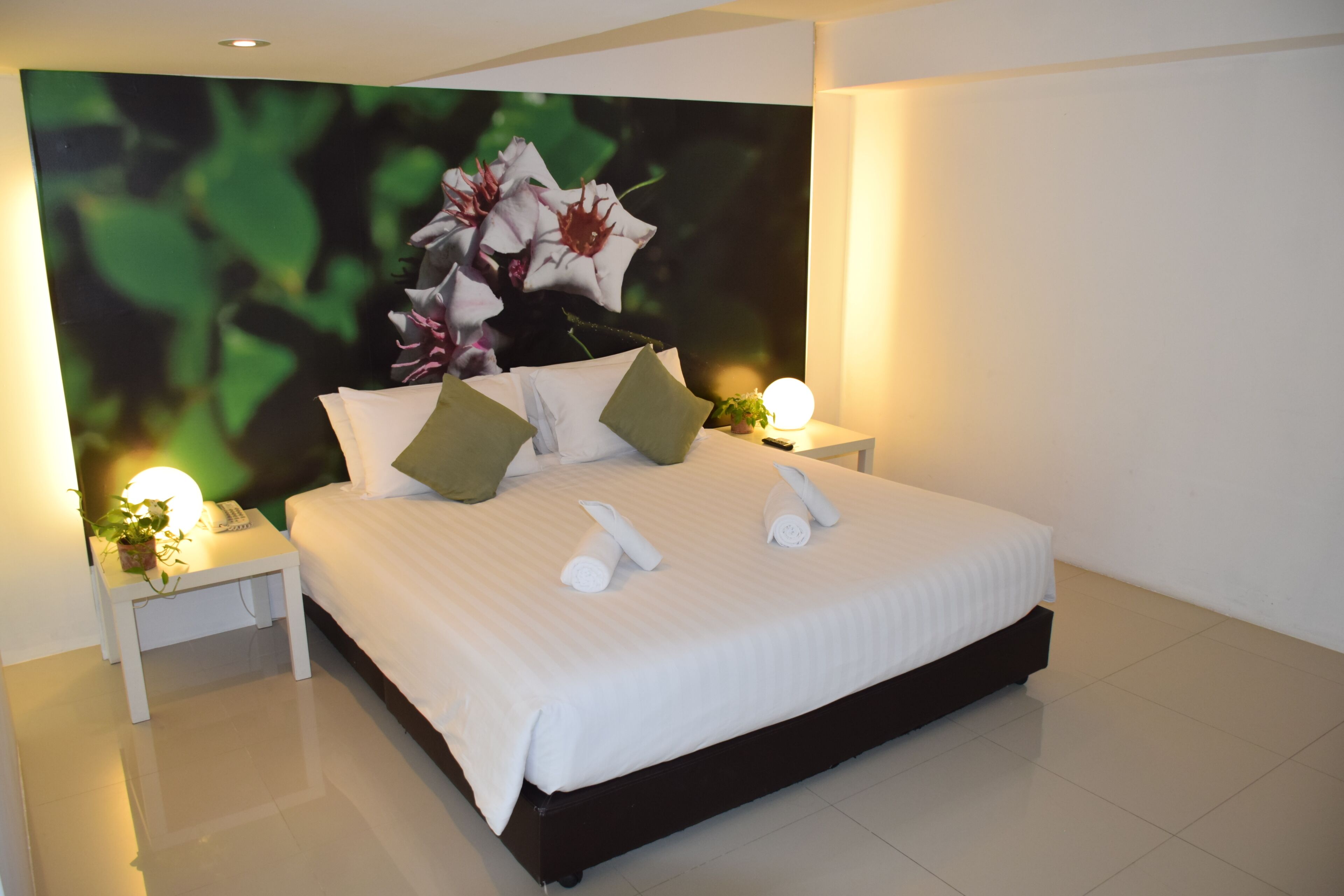 A modern hotel room with a large bed, two bedside tables, a nightstand, a wall-mounted TV, and a potted plant.