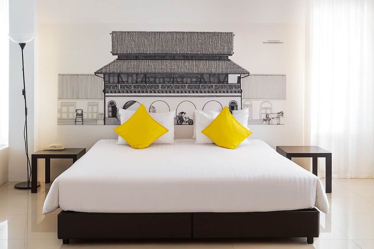 Une chambre d’hôtel avec un grand lit, deux tables de chevet, une lampe sur pied et une fresque murale représentant un bâtiment traditionnel.