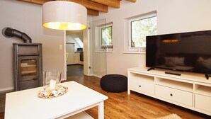 A modern living room with a wooden ceiling, a white table with a candle, a black ottoman, a TV on a white stand, and a kitchen visible through an open door. - Ruhig und Idyllische Lage im Herzen der Insel Rügen - Gutshaus Silvitz Fewo 03 (Bergen auf Rügen)