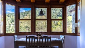 Un comedor con mesa y sillas de madera, ventanas amplias con vista a un bosque y una lámpara colgante.