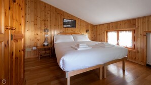 A wooden-paneled room with a large bed, a nightstand, a wall-mounted lamp, and a window with curtains. - Leopardi 3 (Bormio)