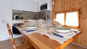 Una mesa de comedor de madera con platos, vasos y cubiertos en una cocina moderna con armarios blancos y una ventana con cortinas.