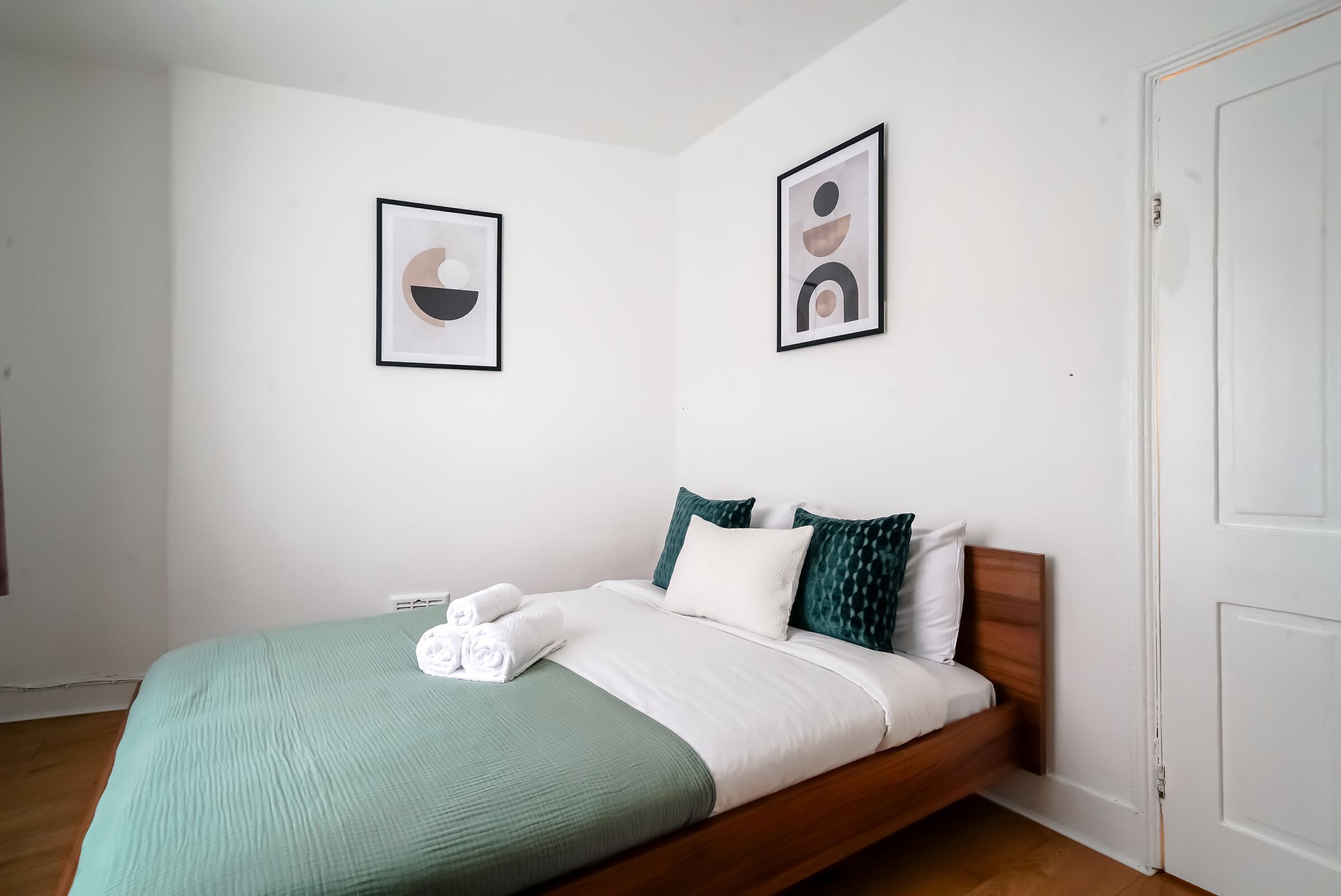 Un lit simple avec une couvre-lit verte et blanche, deux oreillers et une tête de lit en bois. Deux œuvres d’art encadrées sont accrochées au mur.
