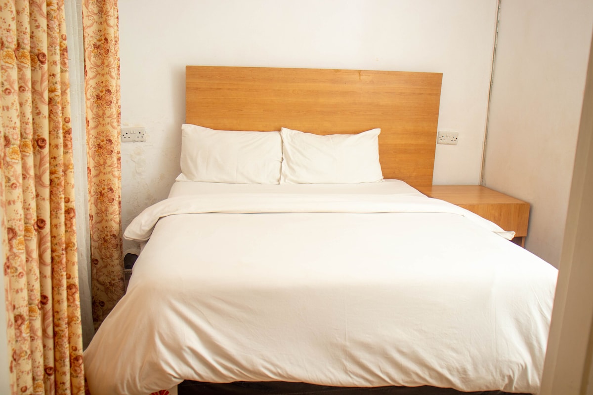 A neatly made bed with a wooden headboard, white bedding, and a floral curtain partially drawn to the side.