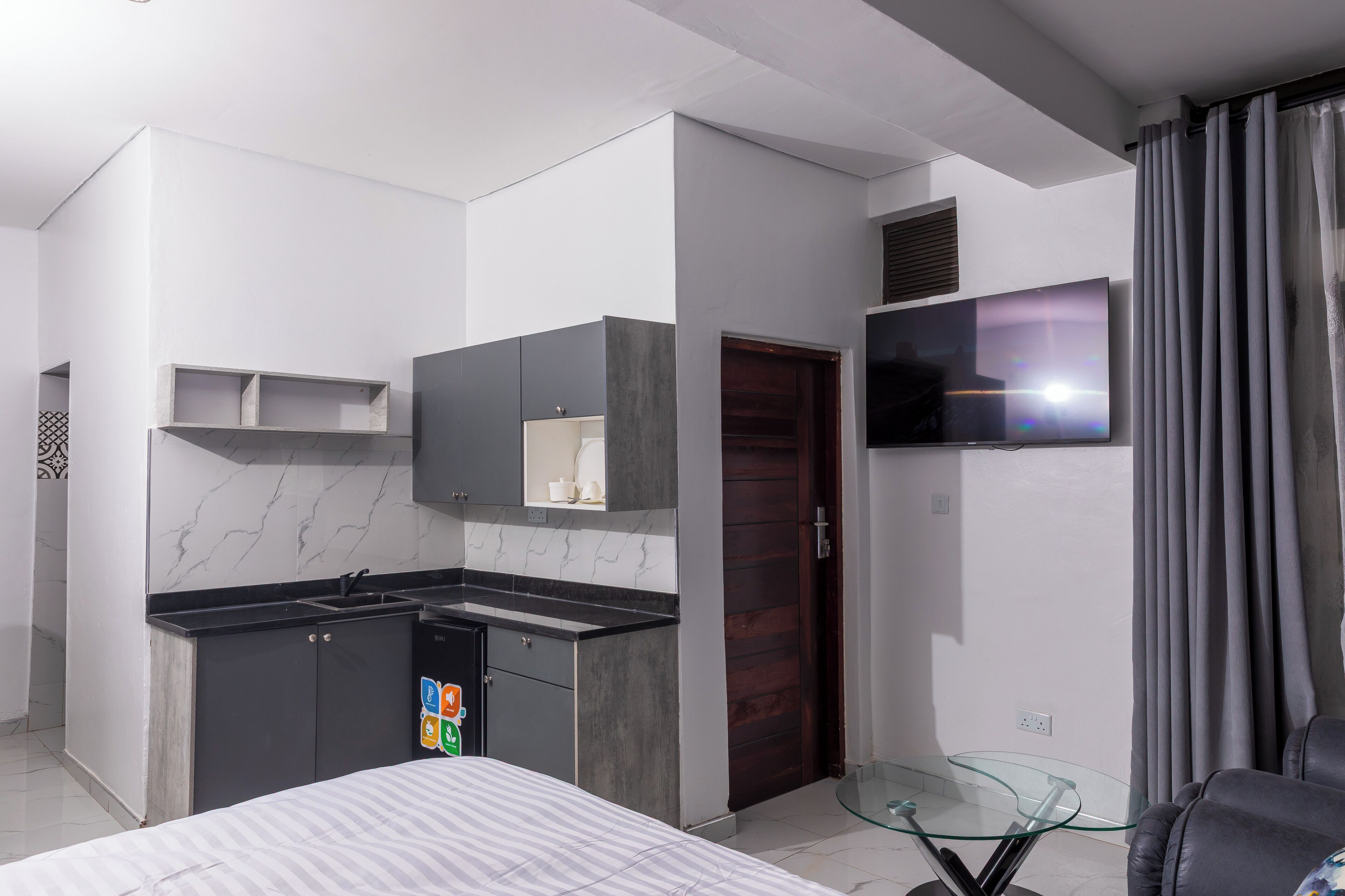 A modern hotel room with a bed, a small kitchenette, a TV mounted on the wall, and a glass table.