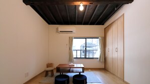 Um quarto com piso de madeira, uma mesa com almofada, dois banquinhos, uma janela com cortinas e um guarda-roupa.