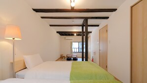 Quarto de hotel com duas camas, uma luminária, um banquinho e uma janela.