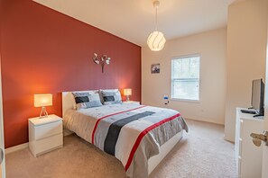 Un dormitorio con cama, mesitas de noche, lámpara, decoración en la pared, ventana y mueble para televisor.