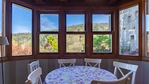 A dining area with a round table covered by a floral tablecloth, surrounded by white chairs, large windows with a view of a forested landscape, and a stone building in the background.