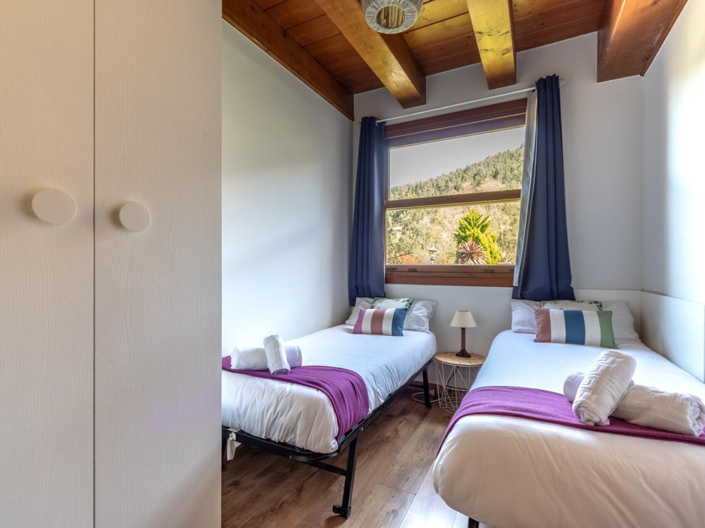 Une chambre avec deux lits, un plancher en bois, une fenêtre avec des rideaux et une vue sur une colline.