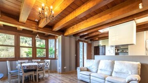 A spacious living room with wooden floors, a white sofa, a dining table with chairs, and large windows offering a view of greenery.