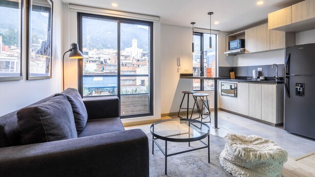 En moderne lejlighed med en sofa, et rundt glasbord, et køkken med et køleskab og udsigt over byen gennem store vinduer.
