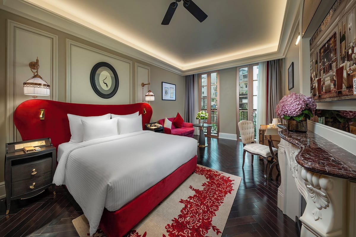 Una habitación de hotel amplia con una cama grande, cabecera roja, un escritorio con silla, una mesita con un jarrón de flores y vistas a la ciudad a través de la ventana.