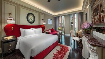 A spacious hotel room with a large bed, a red headboard, a desk with a chair, a small table with a vase of flowers, and a view of the city through the window.