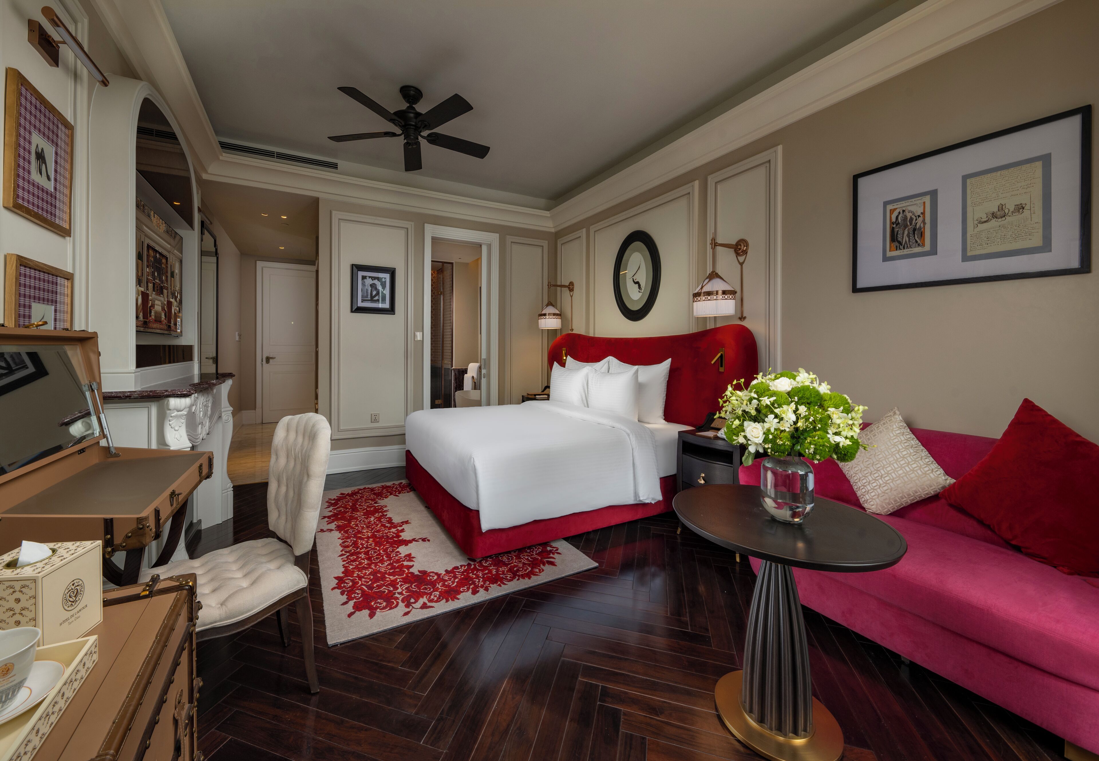 A hotel room with a large bed, a red sofa, a desk with a chair, a small table with a vase, and a ceiling fan.