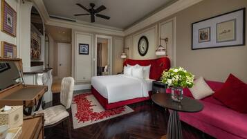 A hotel room with a large bed, a red sofa, a desk with a chair, a small table with a vase, and a ceiling fan.