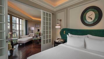 A hotel room with a large bed, a green upholstered headboard, a round mirror, and a view through a glass door to a balcony.