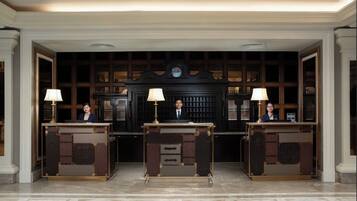 A reception area with three desks, each with a lamp and a person seated behind it. The background features a dark wooden panel with a decorative emblem.