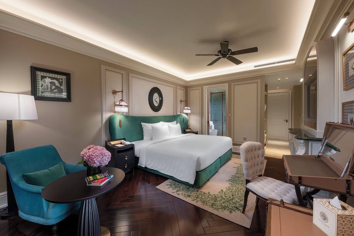 Habitación de hotel con cama, escritorio, silla, mesita de noche, lámpara, ventilador de techo y espejo.