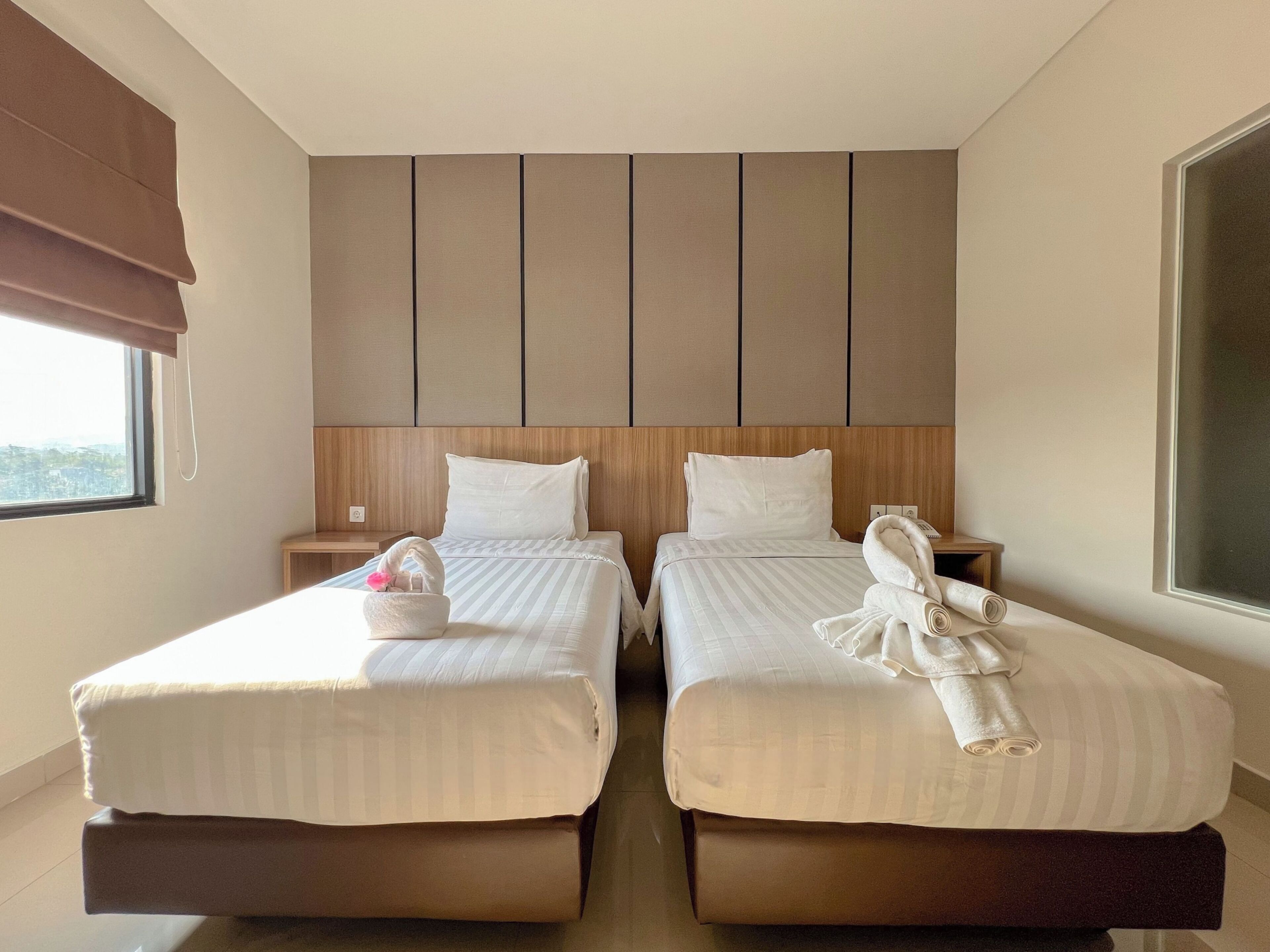 A hotel room with two beds, a wooden headboard, a nightstand, and a window with a view.