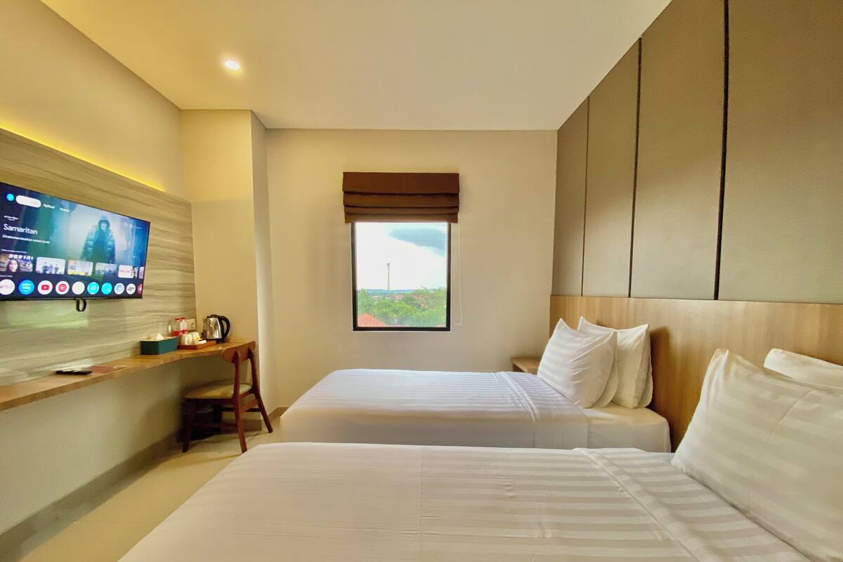 A hotel room with two beds, a TV mounted on the wall, a window with a view, and a wooden headboard.
