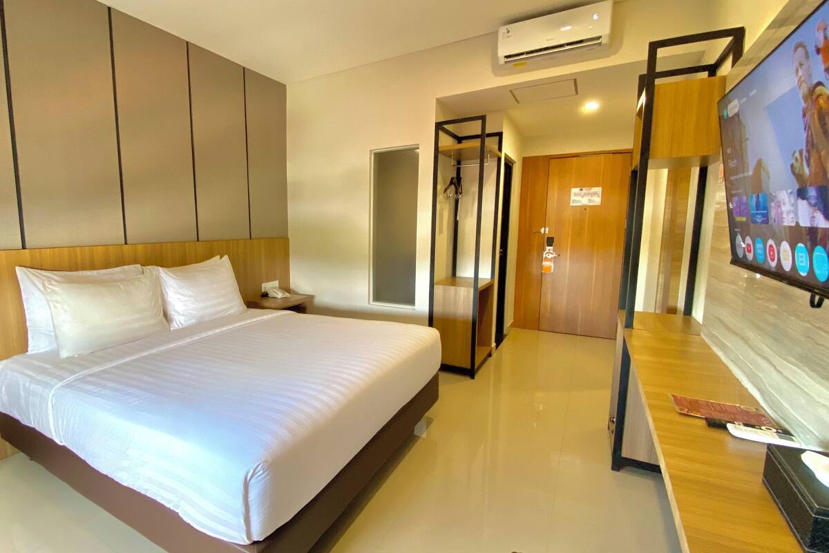 A hotel room with a large bed, a wooden headboard, a bedside table with a phone, a TV on the right, and a door leading to another room.