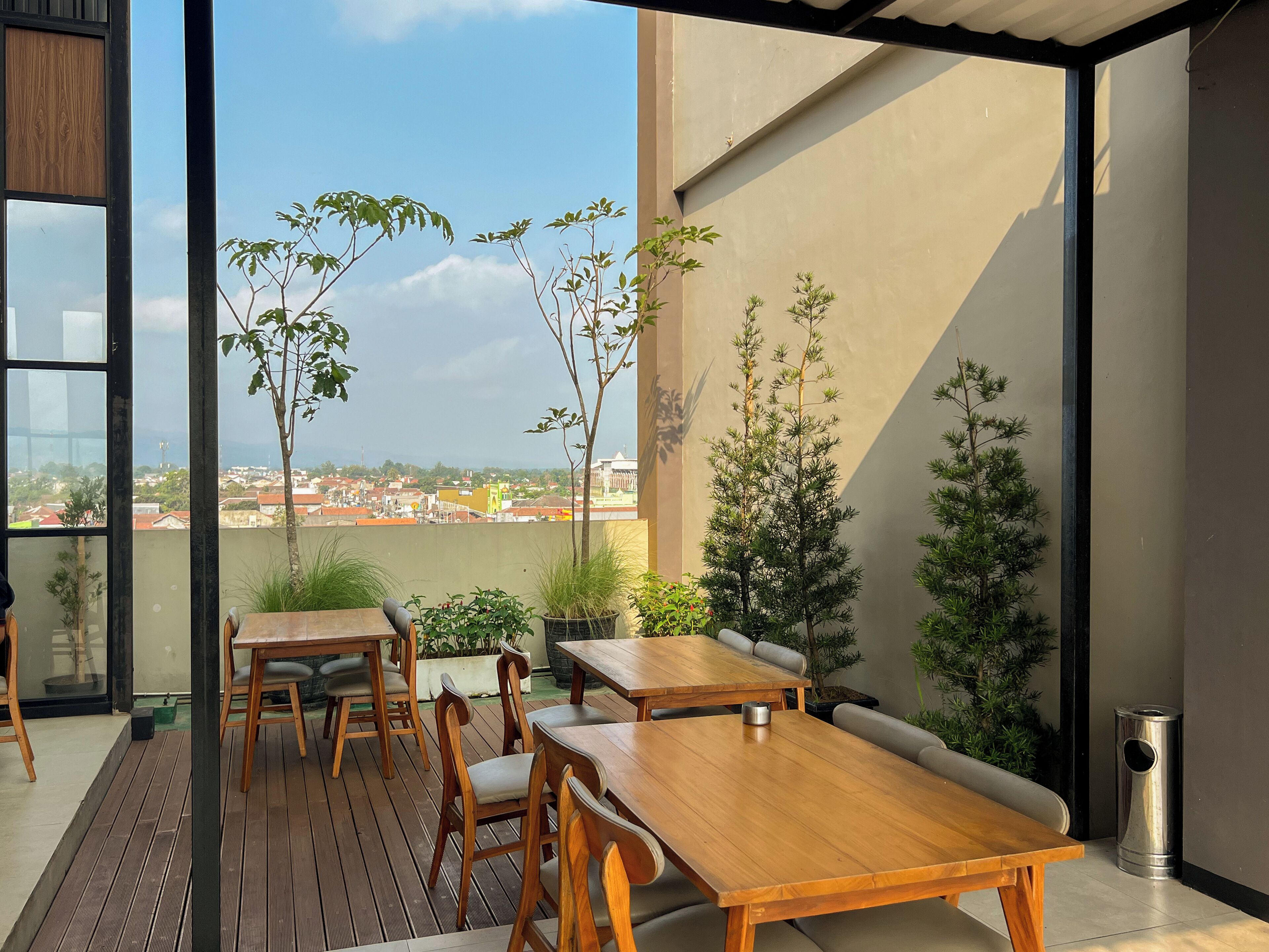 An outdoor dining area with wooden tables and chairs, surrounded by potted plants and a view of the cityscape.