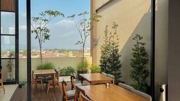 An outdoor dining area with wooden tables and chairs, surrounded by potted plants and a view of the cityscape.
