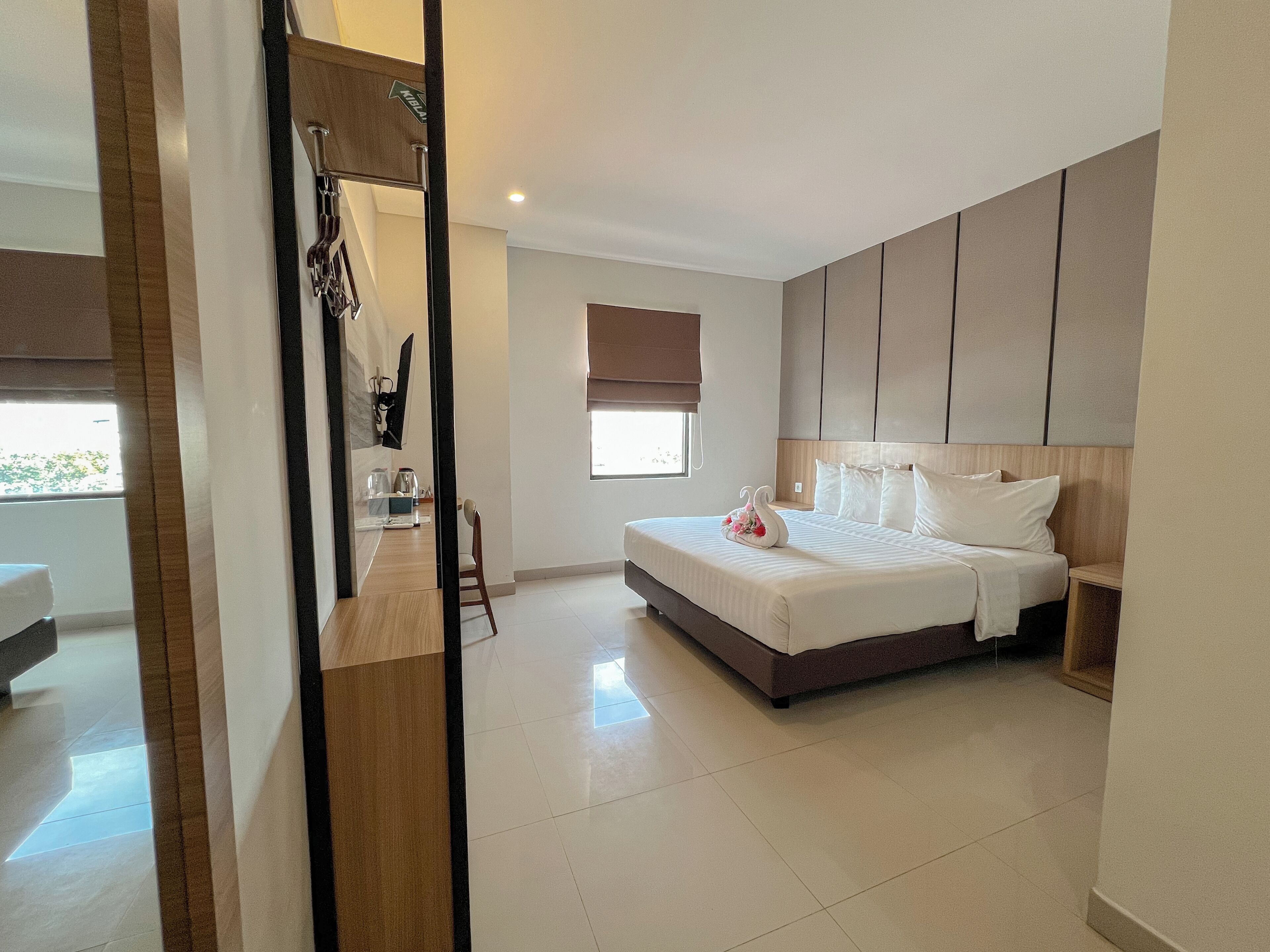 A modern hotel room with a large bed, a bedside table, a chair, a window with a view, and a mirror.
