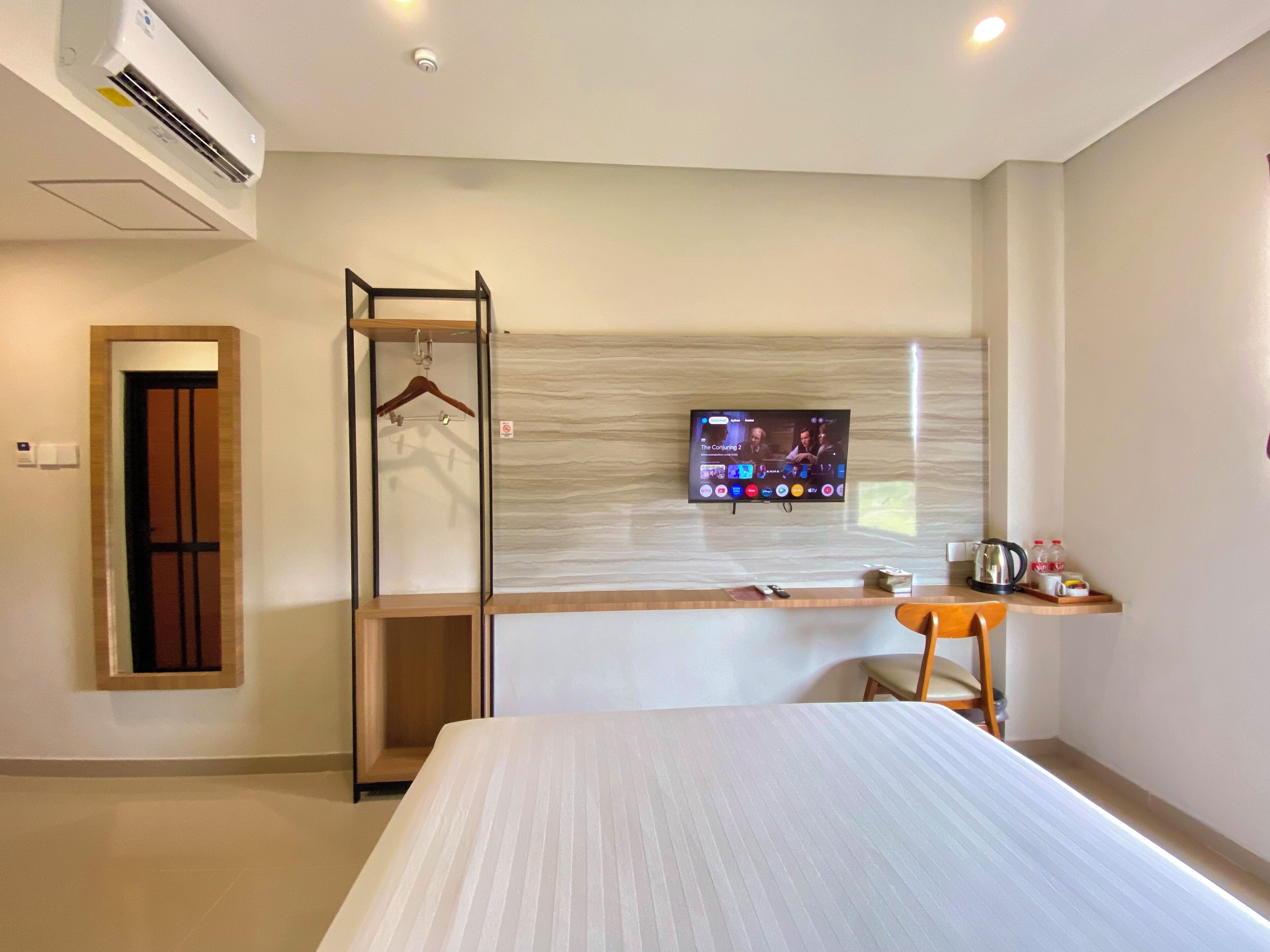 Habitación de hotel con una cama, un televisor montado en la pared, una silla de madera, una mesita con un hervidor y un equipo de aire acondicionado.