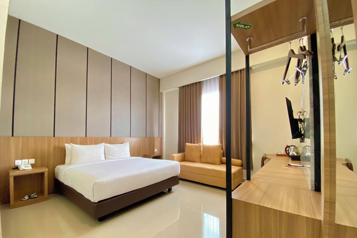 A modern hotel room with a large bed, a sofa, a desk with a chair, and a mirror.
