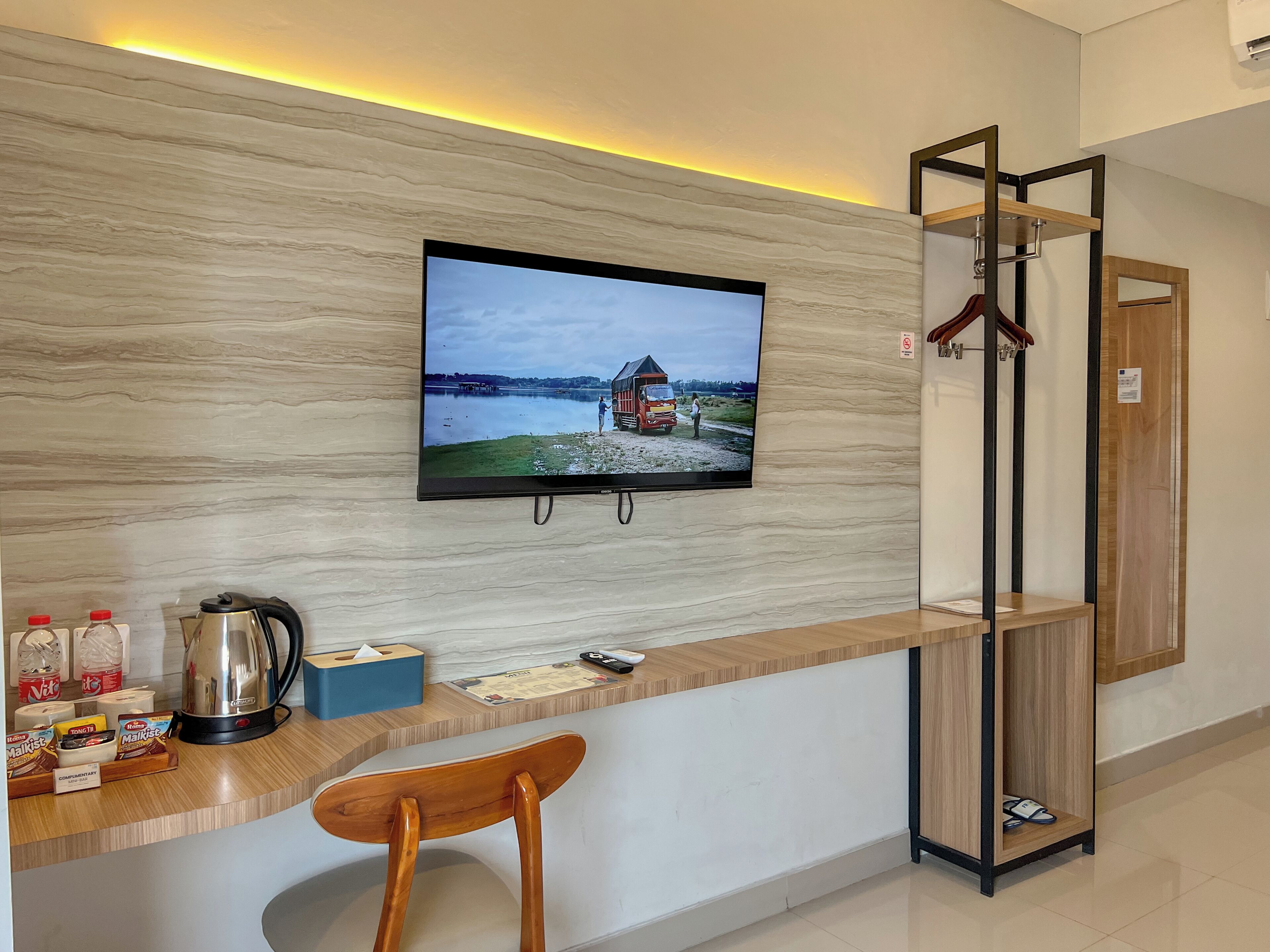 Zona de recepción de hotel con un televisor de pantalla plana montado en una pared de mármol, un mostrador de madera, una silla y un perchero.