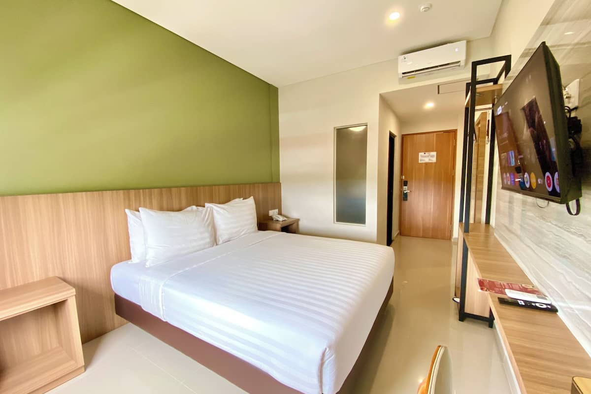 A hotel room with a large bed, a wooden headboard, a desk with a flat-screen TV, and an air conditioning unit on the wall.