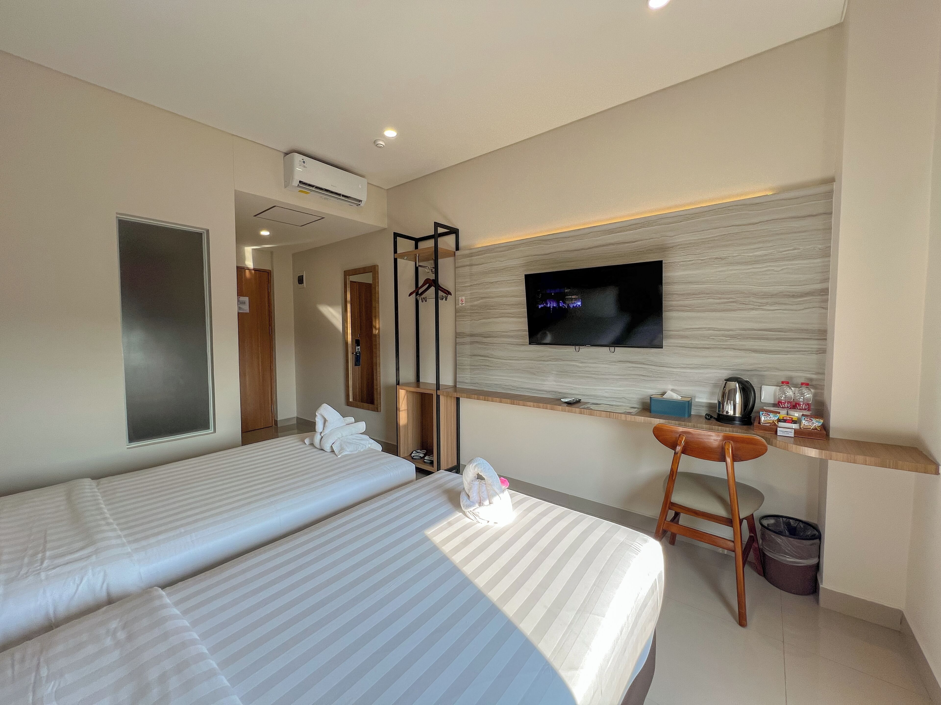 A hotel room with two beds, a TV mounted on the wall, a desk with a kettle, and a trash bin.