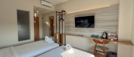 A hotel room with two beds, a TV mounted on the wall, a desk with a kettle, and a trash bin.