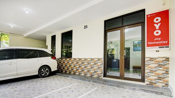 L’entrée d’un hôtel, avec une voiture blanche garée dans l’allée, un panneau portant le numéro 23 au-dessus de la porte et une enseigne rouge faisant de la publicité pour « OYO Joy Homestay ».