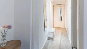 A hallway with a wooden bench, a vase with flowers, and a mirror reflecting a door and a light fixture.